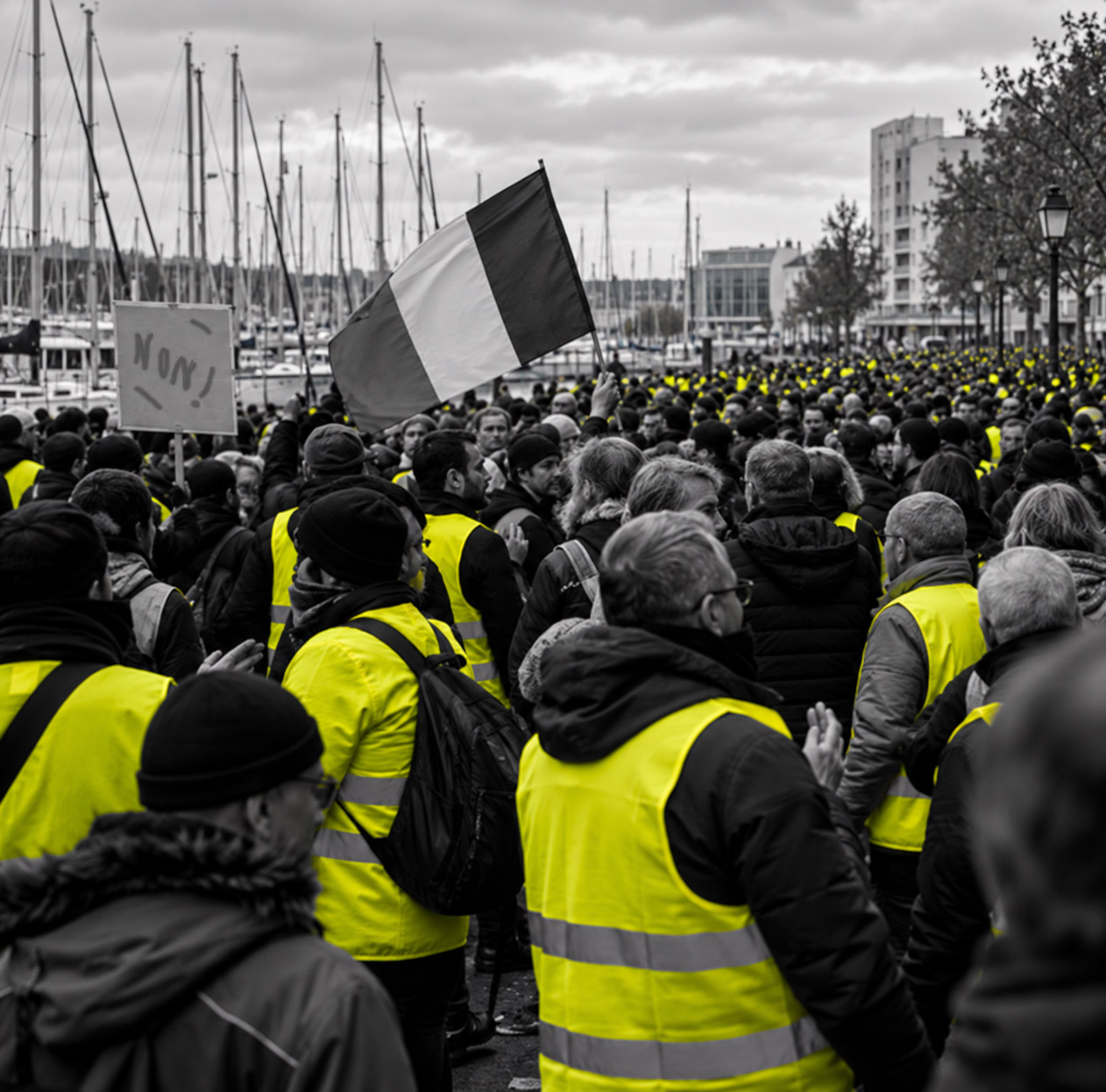 Cortège de gilets jaunes le long d'un port de plaisance breton, vu de l'intérieur du mouvement, traitement éditorial en niveaux de gris avec les gilets en couleur.